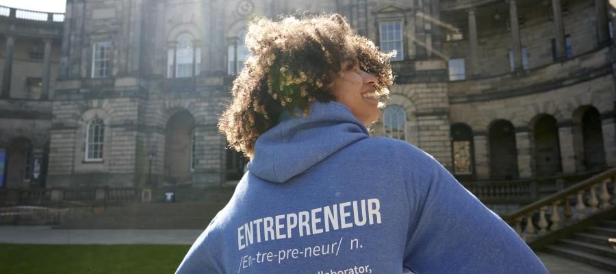 A women wearing a blue hoodie, branded with the word 'entrepreneur' on the back. Standing in front of Old College with the sun shining on her.