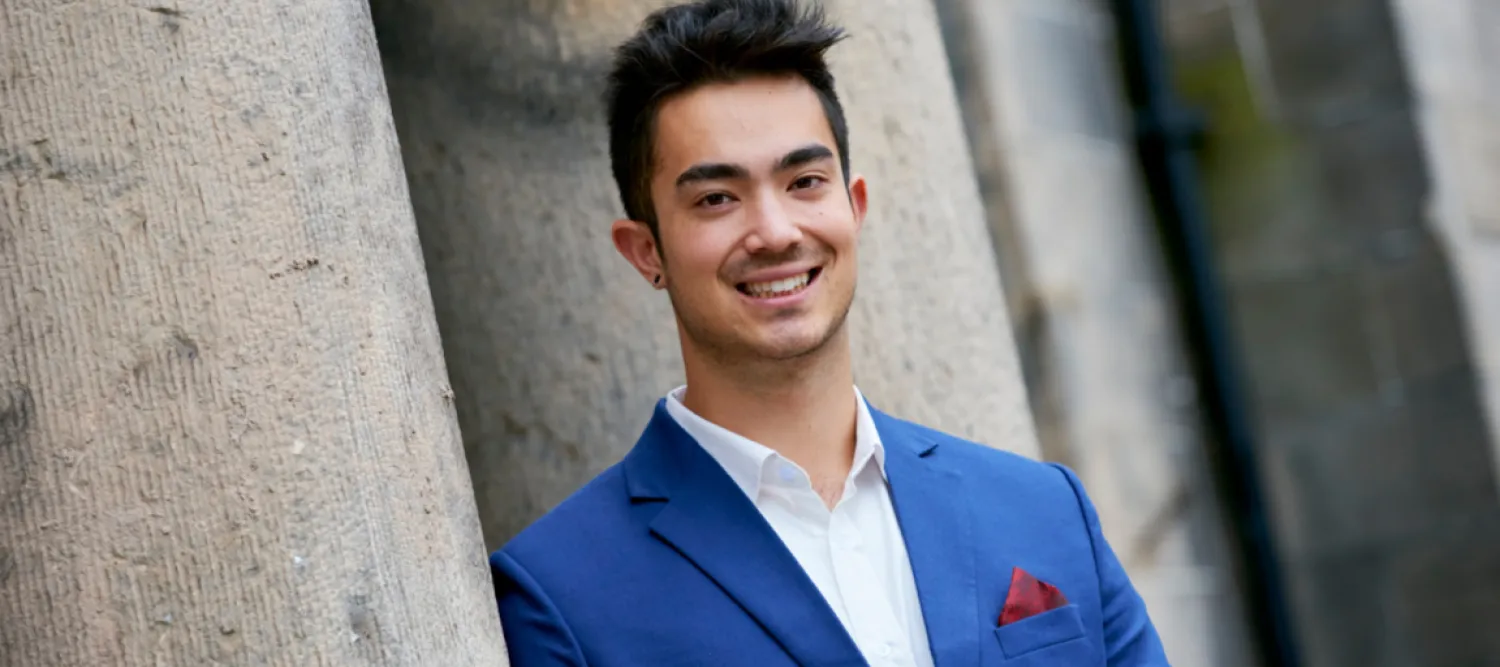 Man wearing suite, smiling at camera. Stone pillars behind him. 