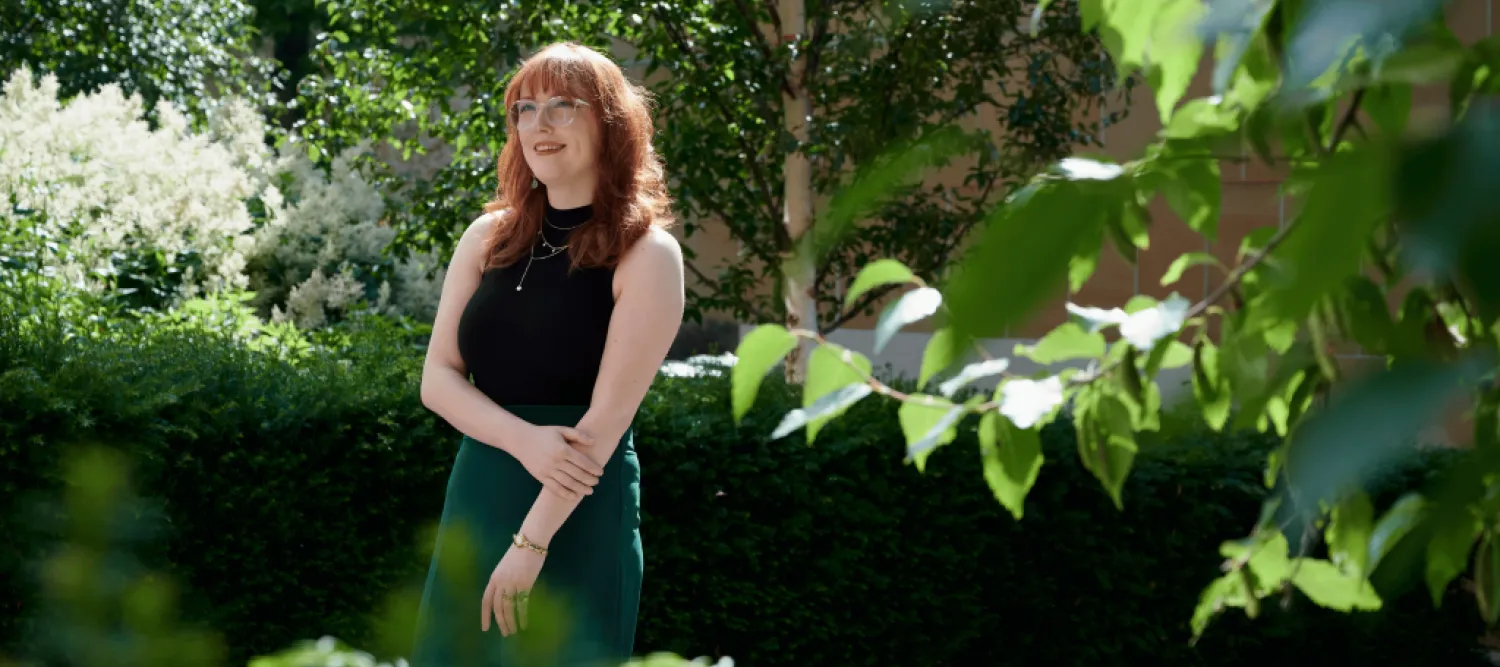 Women with red hair and glasses standing amongst greenery. 