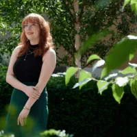 Women with red hair and glasses standing amongst greenery. 