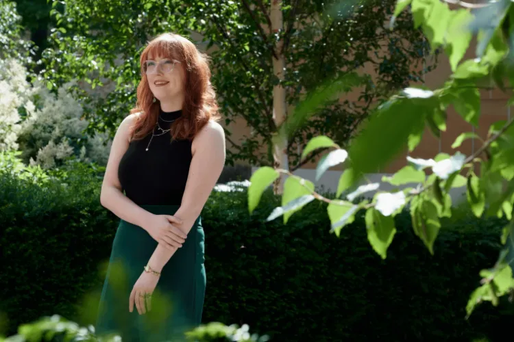 Women with red hair and glasses standing amongst greenery. 