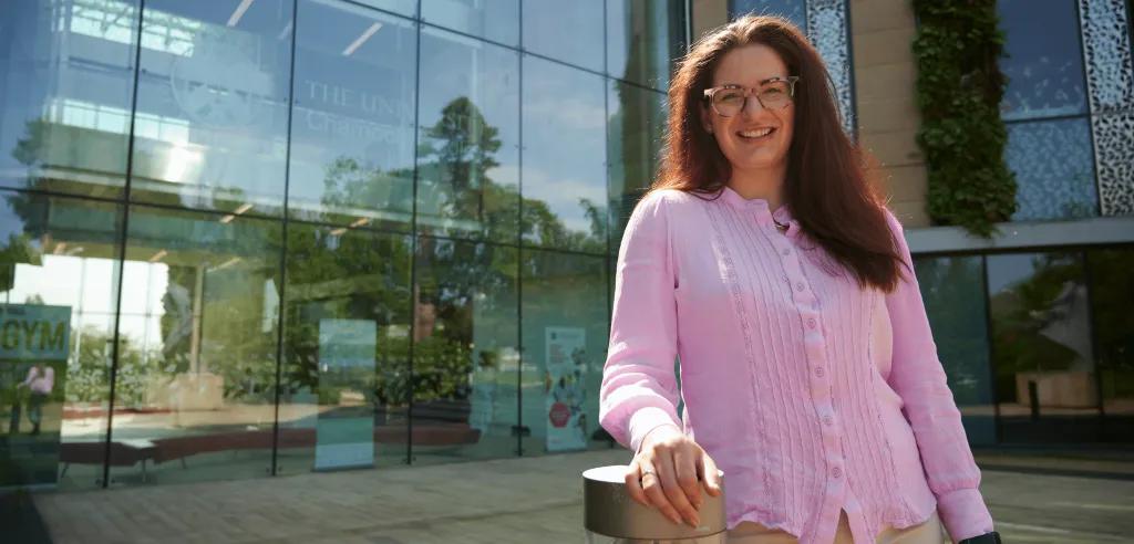 Woman smiling, wearing pink shirt and standing outside University of Edinburgh building.