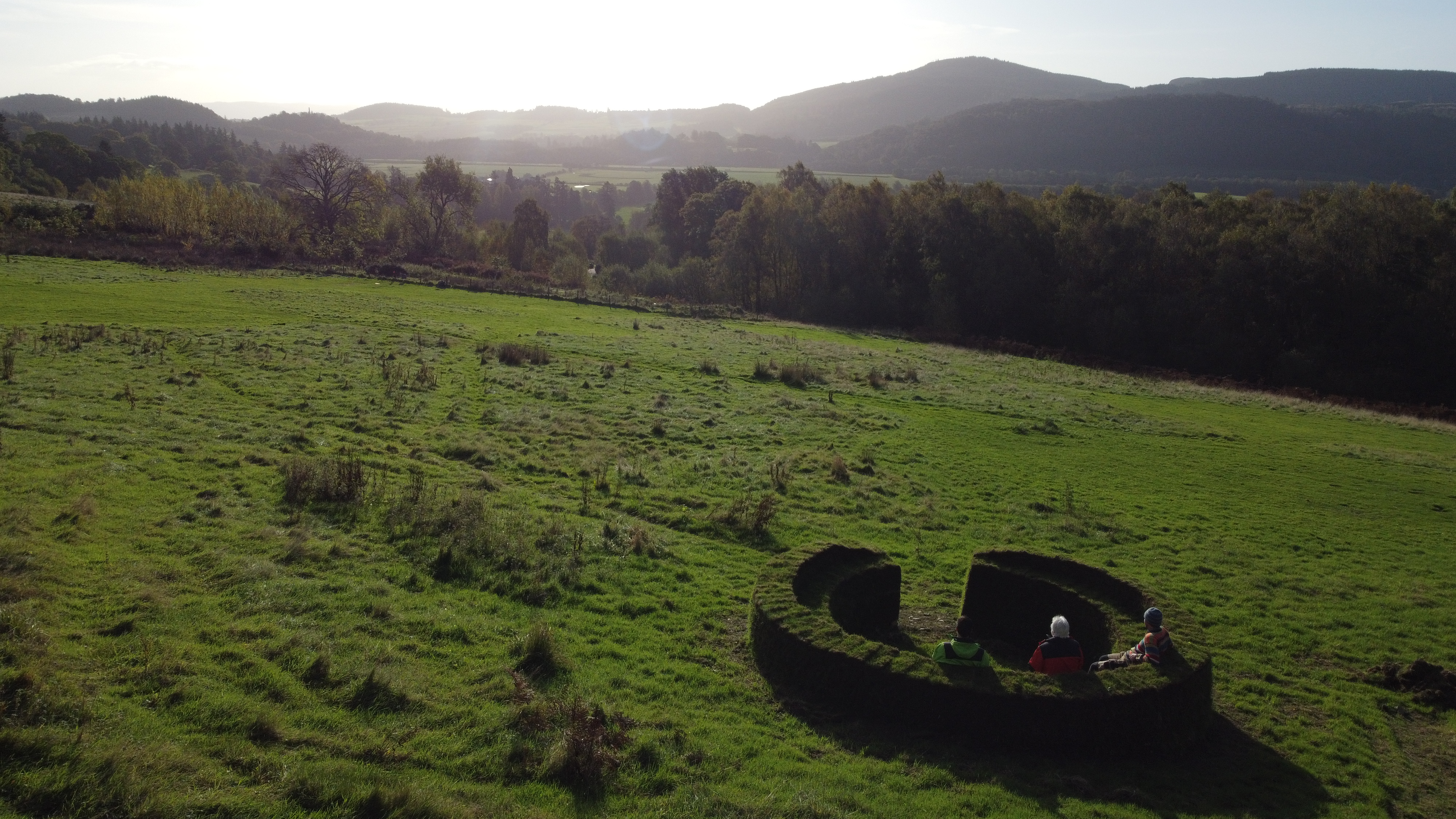 Image of turf bench with people sitting wearing hard hats. Fields, trees and mountains form the backdrop of this image