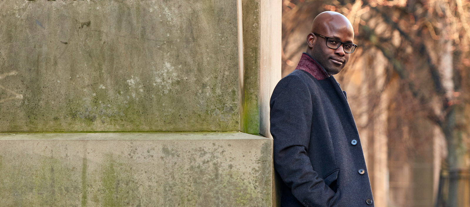 Image of Gbenga Ibikunle standing outside, leaning against an old monument with a tree in the background