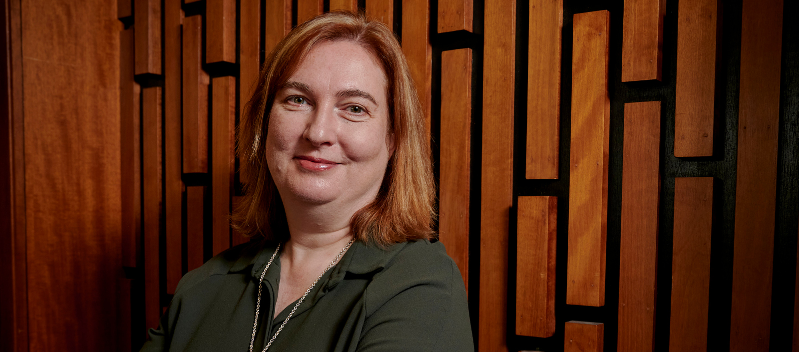 Melissa Terras smiling and standing in front of a wooden wall with a vertical brick effect