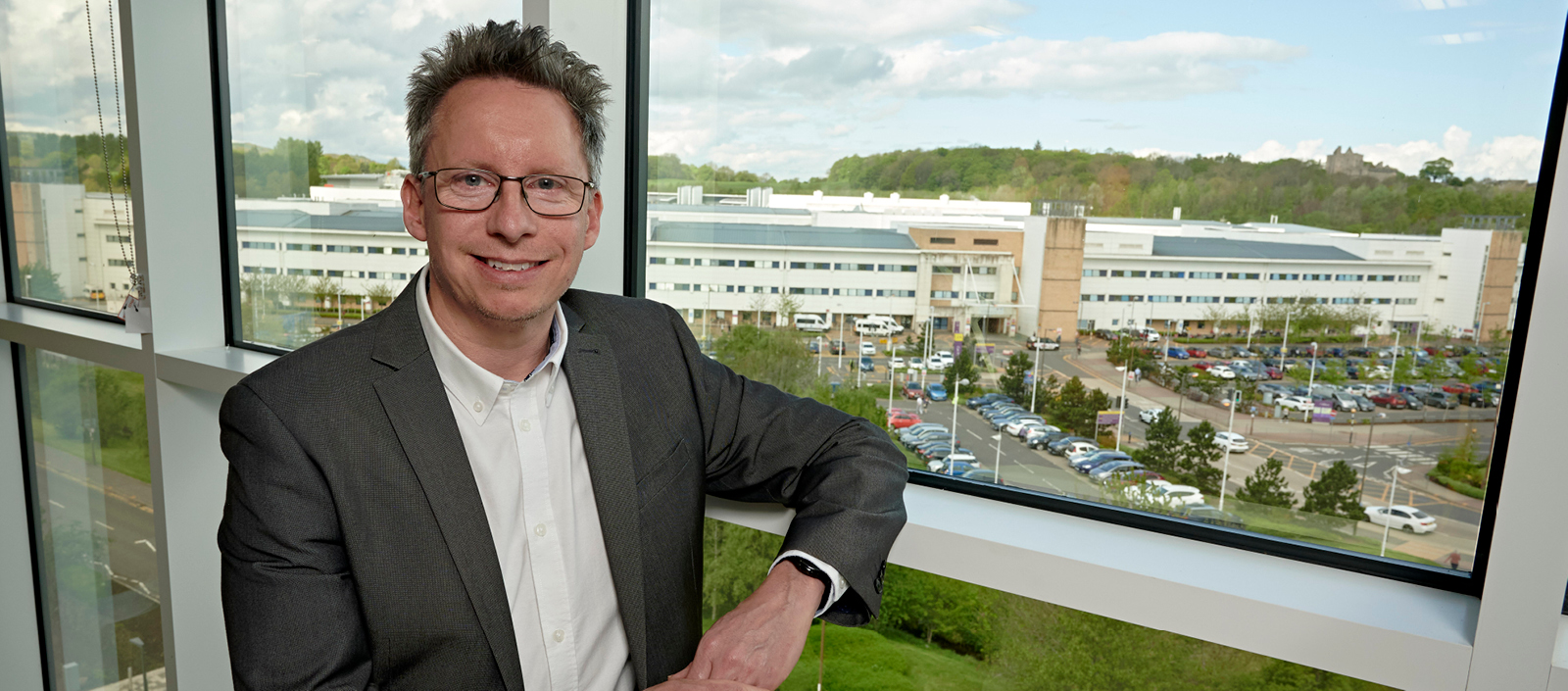 Image of Jonathan Fallowfield wearing a white shirt and grey suit, leaning against a window with University buildings in the background